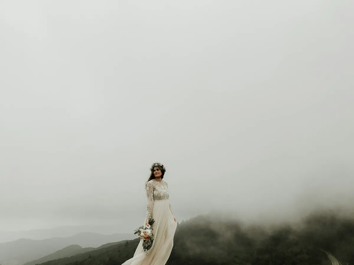 A bride in a long-sleeved lace and chiffon gown standing on a foggy mountain hilltop, her skirt caught by the wind